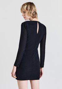 Schwarzes langärmliges Kleid mit taillierter Form, das ein strukturiertes Muster aus kleinen Punkten und ein Schlüsselaugen-Detail am Rücken aufweist.