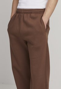 Pantalons de survêtement en coton marron avec taille élastique, poches latérales et coupe décontractée, présentant une texture lisse et une couleur unie.