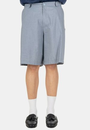Uomo che indossa pantaloncini grigio chiaro fino al ginocchio, calzini bianchi e mocassini neri con nappine, in piedi su uno sfondo semplice.