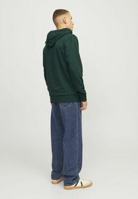 Felpa verde scuro con tasca a marsupio, indossata con jeans denim blu larghi e sneakers crema con una striscia verde. Tessuto morbido, vestibilità casual.