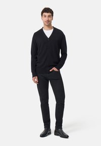 Černý cardigan s výstřihem do V, čtyřmi knoflíky a slim fit střihem, v kombinaci s černými džíny a černými kotníkovými botami na modelce proti jednoduchému pozadí.
