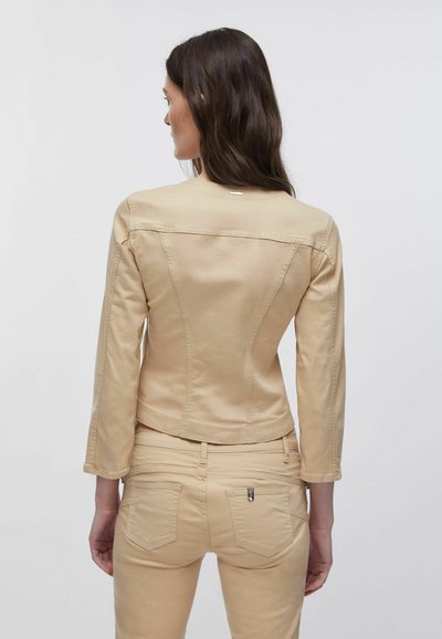 Femme aux longs cheveux bruns vue de dos, portant une veste beige ajustée et un pantalon assorti sur un fond clair uni.