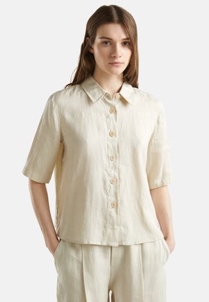 Vrouw draagt een beige korte mouwen overhemd met kraag en bijpassende beige broek, staand met handen in de zakken.