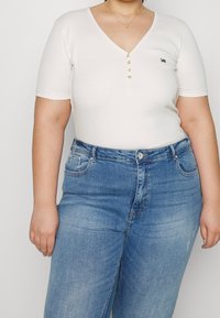 Bílý přerušovaný top s výstřihem do V a krátkými rukávy, který je ozdoben řadou zlatých knoflíků, v kombinaci s modrými denimovými džínami se světlým efektem a kapsami.