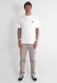 T-shirt en coton blanc avec un petit graphisme bleu sur la poitrine, associé à un pantalon beige et des baskets noires. Le modèle est debout.