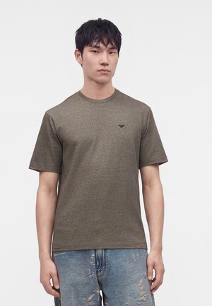 Ein junger Mann trägt ein strukturiertes olivgrünes T-Shirt mit kurzen Ärmeln und ausgewaschene blaue Jeans und steht vor einem schlichten weißen Hintergrund.