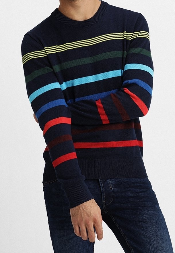Homme portant un pull bleu marine à rayures horizontales jaunes, vertes, bleues et rouges, associé à un jean bleu foncé, les bras croisés.