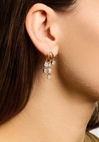 Pendientes de tono dorado con cristales iridiscentes en cascada de varios tamaños. El diseño incluye tres racimos colgando de un aro minimalista.