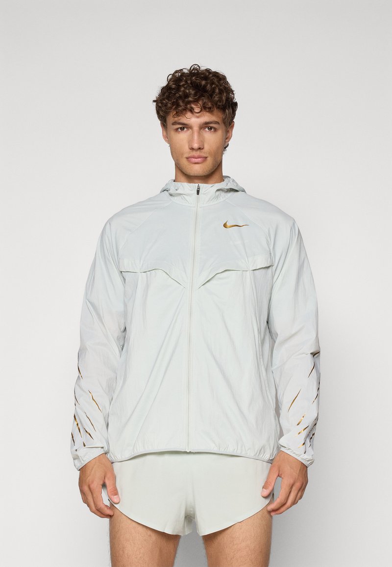 Veste Nike gris clair avec fermeture éclair sur le devant, capuche et logo doré ; possède des manches raglan et des détails de coupe subtils sur les bras.