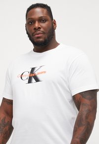 Hvit t-skjorte i bomull med en svart og oransje "Calvin Klein Jeans" logo, korte ermer, rund halsutringning, med synlige arm-tatoveringsdetaljer.