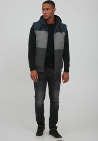 Kapuze Weste mit einem Farbblockdesign in Dunkelblau, Grau und Hellgrau. Zwei Seitentaschen und ein strukturiertes Strickpullover darunter.