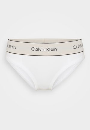 White cotton brief with a beige elastic waistband featuring black "Calvin Klein" text. Smooth texture, mid-rise design, and classic shape.