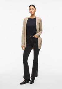 Beige cardigan over a svart topp, kombinert med svarte vide jeanstype bukser og svarte ankelstøvler. Cardiganen har åpen front og ribbestrikkede ermer.