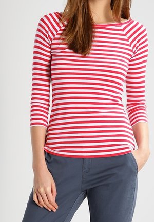 Femme portant un t-shirt à manches longues rayé horizontalement rouge et blanc avec des manches trois-quarts et un pantalon gris foncé, main dans la poche.