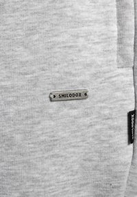 Grauer Sweatshirtstoff mit einer weichen Textur. Verfügt über ein Metallschilder mit der Aufschrift "SMILODOX" und ein kleines schwarzes Etikett an der Seite.