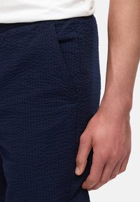 Granatowe spodnie z teksturowanej bawełny z kieszenią z boku. Materiał ma wzór przypominający gofry, co dodaje zainteresowania wizualnego i głębi.