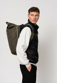 Lo zaino presenta un'esterno verde oliva con texture, spallacci regolabili e un design snello. Il modello indossa un gilet nero sopra un maglione bianco.