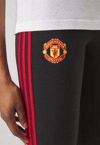 Gros plan sur un pantalon de sport noir avec des bandes rouges et le logo jaune-rouge de Manchester United sur la cuisse, porté avec une chemise blanche.