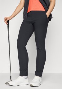 Navy women's golf pants made of stretchy fabric, featuring a slim fit and side pockets, paired with white athletic shoes.