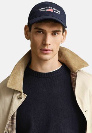 Giovane uomo con cappellino blu navy con la scritta "GANT USA SPORT", maglione blu navy e cappotto beige con colletto ampio, di fronte a uno sfondo uniforme.