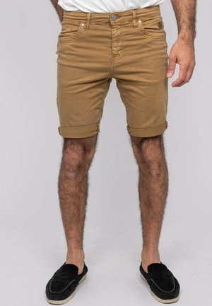 Homme portant un short marron retroussé, une chemise blanche et des chaussures noires sans lacets, debout devant un fond clair et uni.