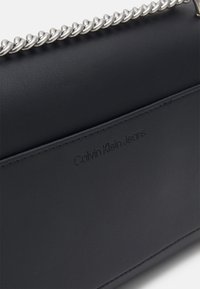 Czarna skórzana torba na ramię z srebrnym łańcuszkiem. Posiada wytłoczone logo "Calvin Klein Jeans" z przodu, gładką teksturę oraz minimalistyczny design.