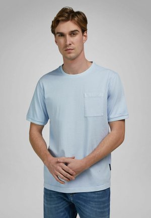 Jonge man met lichtbruin haar, gekleed in een lichtblauw T-shirt met korte mouwen en een borstzak, en een blauwe spijkerbroek, met zijn handen voor zich gevouwen.