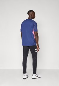 T-shirt de sport bleu marine avec des accents roses et des manches texturées, associé à un pantalon noir avec des logos en chevron blancs et des zip.