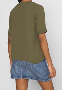 Persona che indossa una maglietta verde oliva a maniche corte e una mini gonna di jeans blu, vista da dietro su uno sfondo chiaro e semplice.