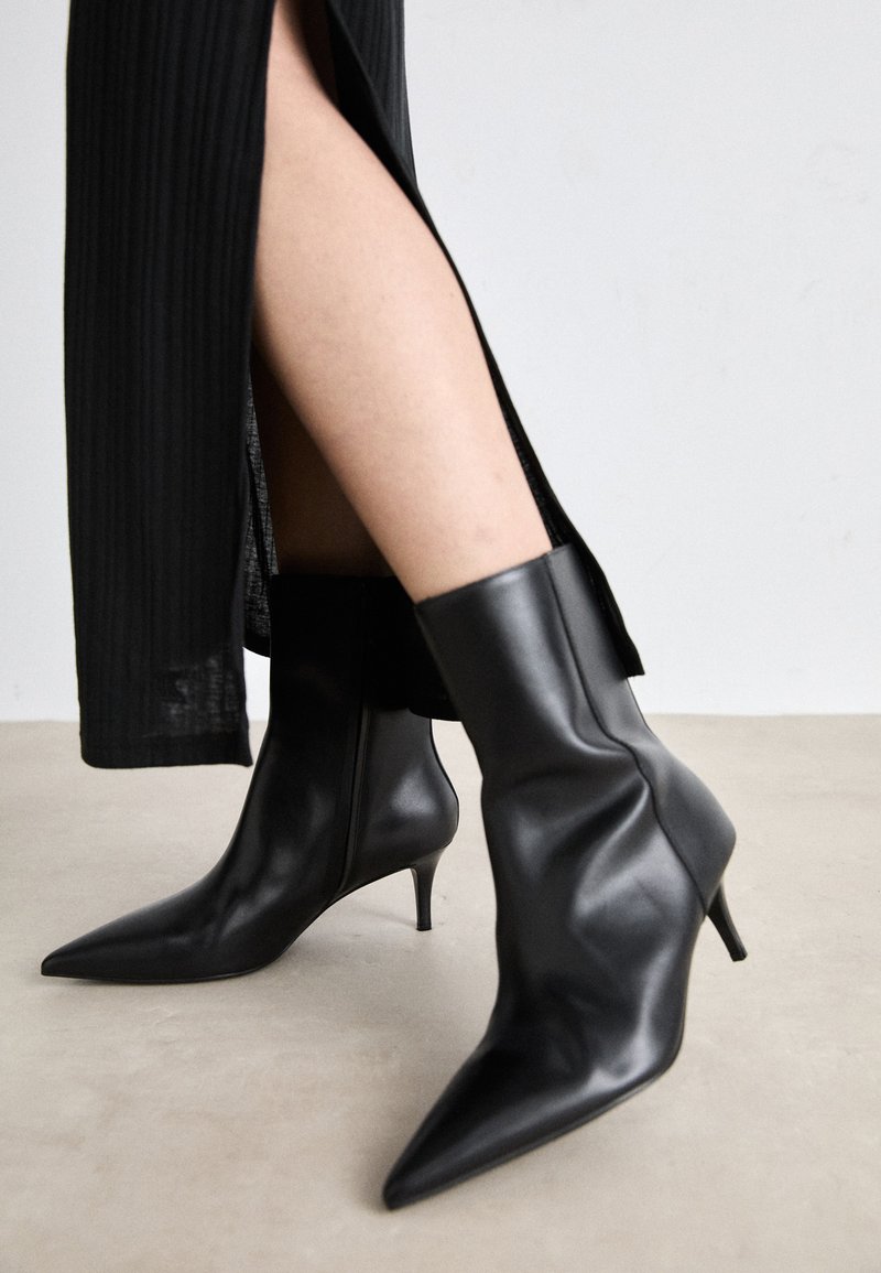 Botins de couro pretos até ao tornozelo com biqueira pontiaguda e salto fino, apresentando uma textura suave e um design ajustado ao tornozelo, emparelhados com uma saia preta com pregas.