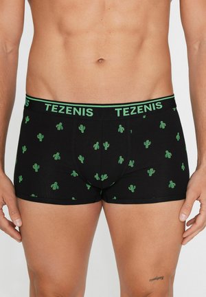 Mężczyzna noszący czarne bokserki z małym zielonym wzorem kaktusa i pasem z logo "TEZENIS", stoi z rozluźnionymi ramionami.