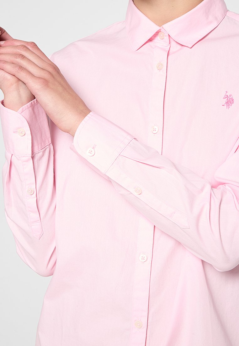 Chemise à manches longues boutonnière de couleur rose clair en coton. Elle présente un col classique, un logo brodé, et des poignets à boutons avec des boutons assortis.