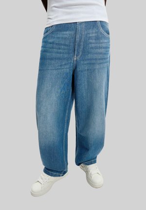 Losse blauwe denim jeans gedragen met witte sneakers en een wit shirt, tegen een effen grijze achtergrond.