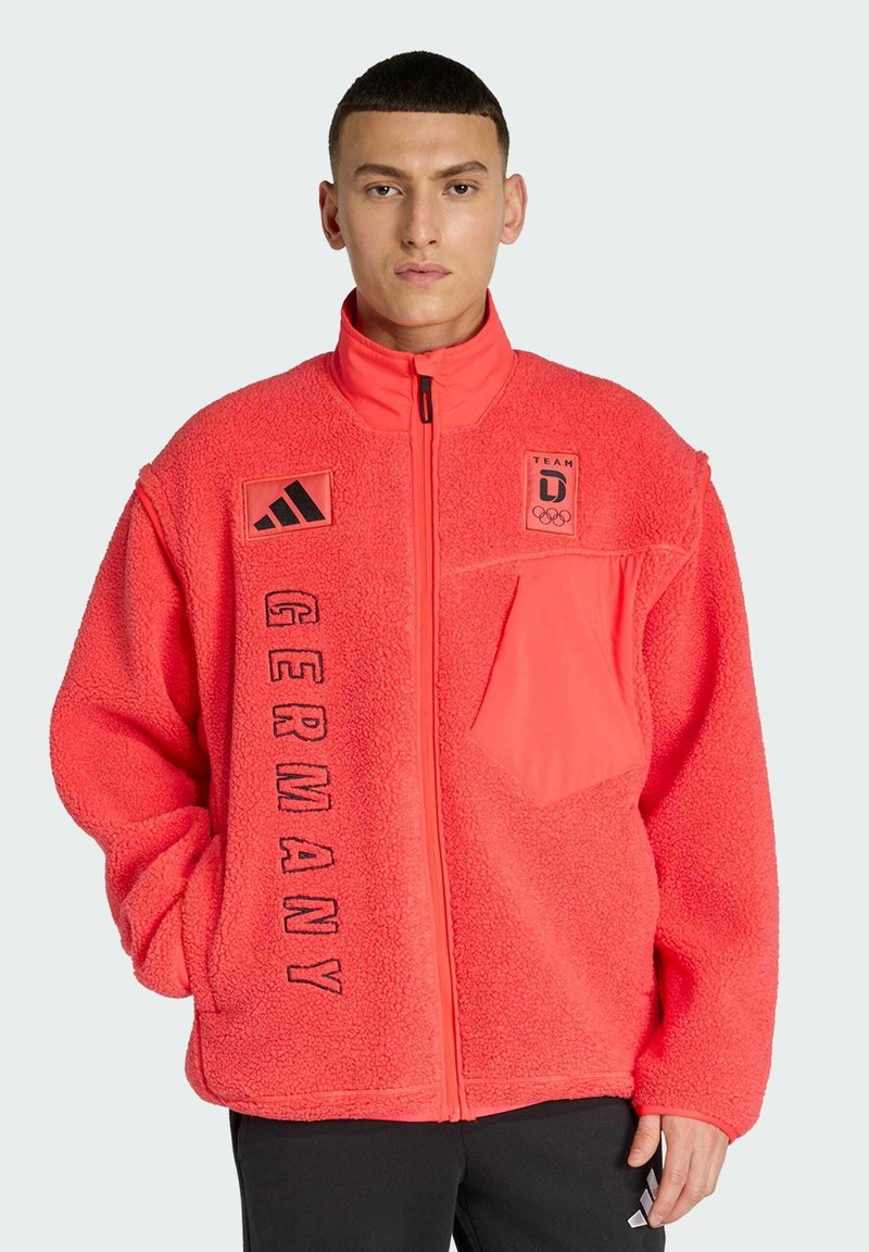 Chaqueta de forro polar de neón rosa con cuello alto, cremallera frontal, texto "GERMANY" y parches con logo en el pecho y la manga. Incluye un bolsillo.