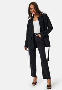 Sort tekstureret jakke med to frontlommer, knaplukning og bånd i taljen, båret over en hvid top og parret med mørke jeans og sandaler.