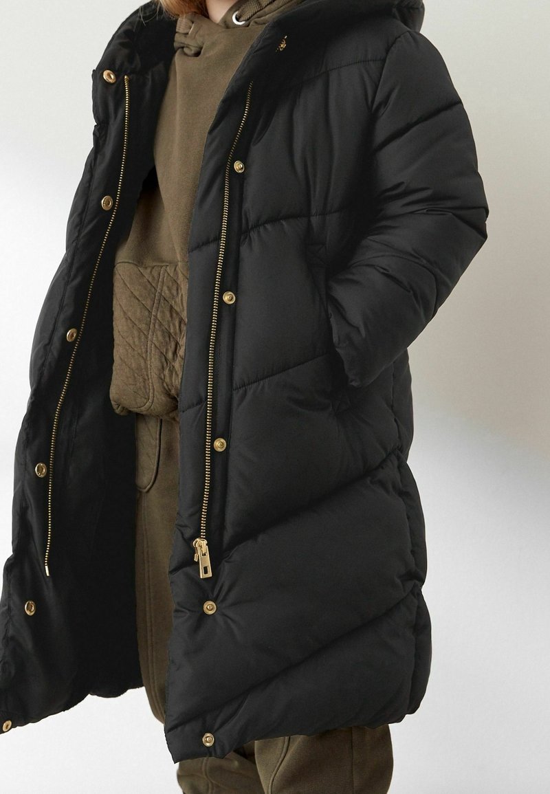Personne portant un manteau matelassé noir avec des boutons et une fermeture éclair dorés, par-dessus un sweat à capuche matelassé vert olive et un pantalon assorti.
