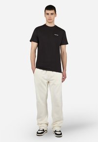 Camiseta de algodón negra con cuello redondo y pequeño logo blanco en el pecho, combinada con pantalones de chándal color crema y zapatillas negras y blancas.