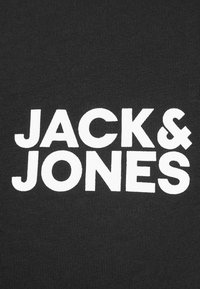 Musta kangas, jossa on poimuttunut rakenne ja rohkeat valkoiset isot kirjaimet, joissa lukee "JACK & JONES." Fontti on moderni ja kaikki kirjaimet ovat isoja.
