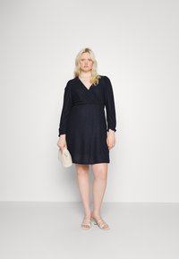 Vestido em tecido azul marinho escuro com padrão texturizado, mangas bufantes longas e decote em V. Combinado com uma clutch bege e sandálias rasas.