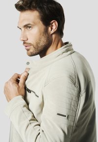 Giacca in pelle beige chiaro con texture trapuntata sulle spalle, tasca con cerniera e dettaglio colletto con bottone a pressione, caratterizzata da un design elegante e moderno.