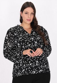 Svart blus med vita abstrakta fläckar, långa ärmar och rynkad halsringning. Bärs med en stor grön ring, mot en neutral bakgrund.