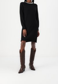 Robe noire en maille à manches longues avec un col décoratif argenté, associée à de hautes bottes de cowboy en cuir marron ornées de motifs en relief.