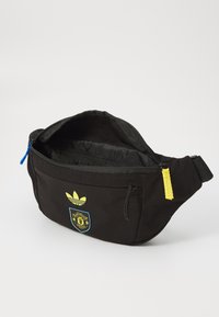 Black fanny pack featuring a yellow Adidas logo and Manchester United emblem, with a zip closure and a blue and yellow zipper pull.