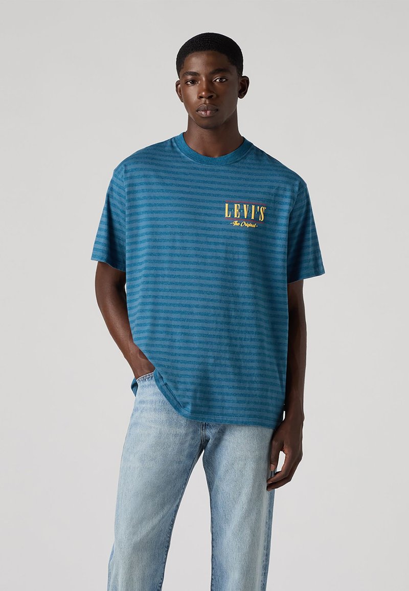 T-shirt de manga curta azul às riscas com decote arredondado e um logo amarelo "LEVI'S" no lado esquerdo do peito. Usado com jeans azul claro.