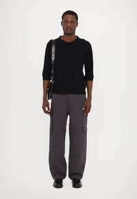 Zwarte gebreide poloshirt met drie knopen, gecombineerd met losse, grijze cargobroek met zakken, en zwarte schoenen.