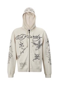 Sudadera beige con cremallera y capucha. Presenta diseños gráficos en negro, incluyendo texto e ilustraciones de una calavera y un águila. Dos bolsillos frontales.