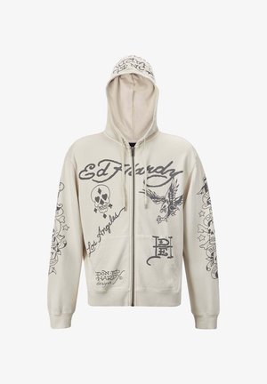 Beige zip-up huppari, jossa on huppu. Siinä on mustia graafisia kuvastoja, mukaan lukien teksti ja kuvastot pääkallosta ja kotkasta. Kaksi etutaskua.