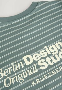 Grün gestreiftes T-Shirt mit einem grafischen Druck in cremefarbener Schrift: "Berlin Design Studio Original Kreuzberg" auf der Vorderseite. Baumwollstoff.