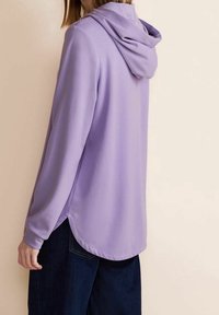 Lila långärmad hoodie med avslappnad passform, utrustad med huva, sidslitsar vid fållen och mjuk tygstruktur. Kombinerad med mörka jeans.