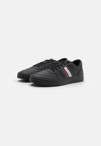 Sneaker sintetiche nere con suola in gomma piatta. Le caratteristiche includono un design con lacci e una striscia a contrasto in rosso, bianco e blu sui lati.